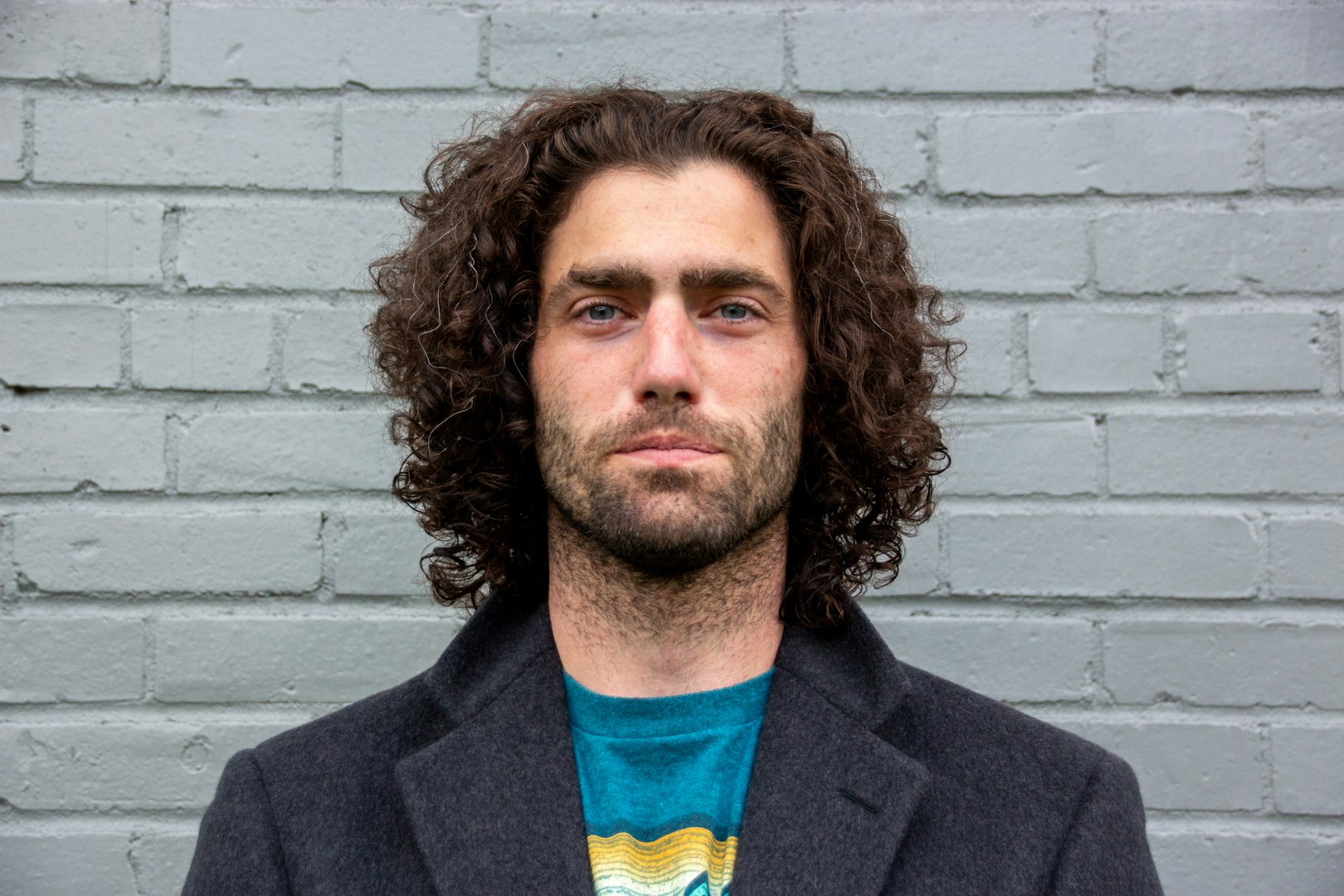A man in a teal work jacket stands in a room with shelves in the background filled with various containers and supplies. He has short, tousled hair and a serious expression.